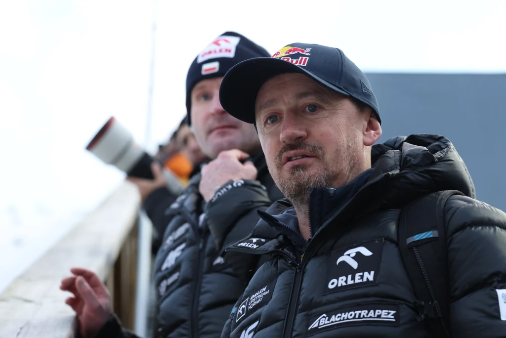 Adam Małysz - prezes Polskiego Związku Narciarskiego. W tle trener kadry polskich skoczków narciarskich - Maciej Maciusiak Dwóch mężczyzn w kurtkach zimowych i czapkach z logo sponsorów, stojących na świeżym powietrzu, jeden z nich patrzy w stronę aparatu, drugi trzyma aparat fotograficzny.