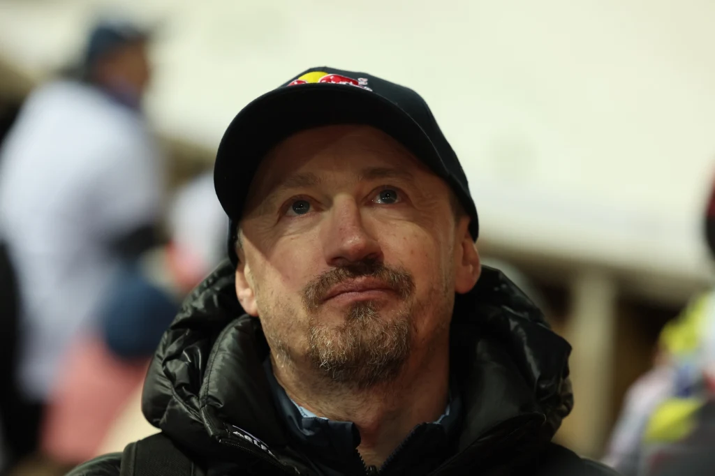 Adam Małysz Mężczyzna w czapce z daszkiem z logo Red Bull, ubrany w ciemną kurtkę, patrzący lekko w górę z zamyśloną miną, rozmyte tło z innymi osobami.