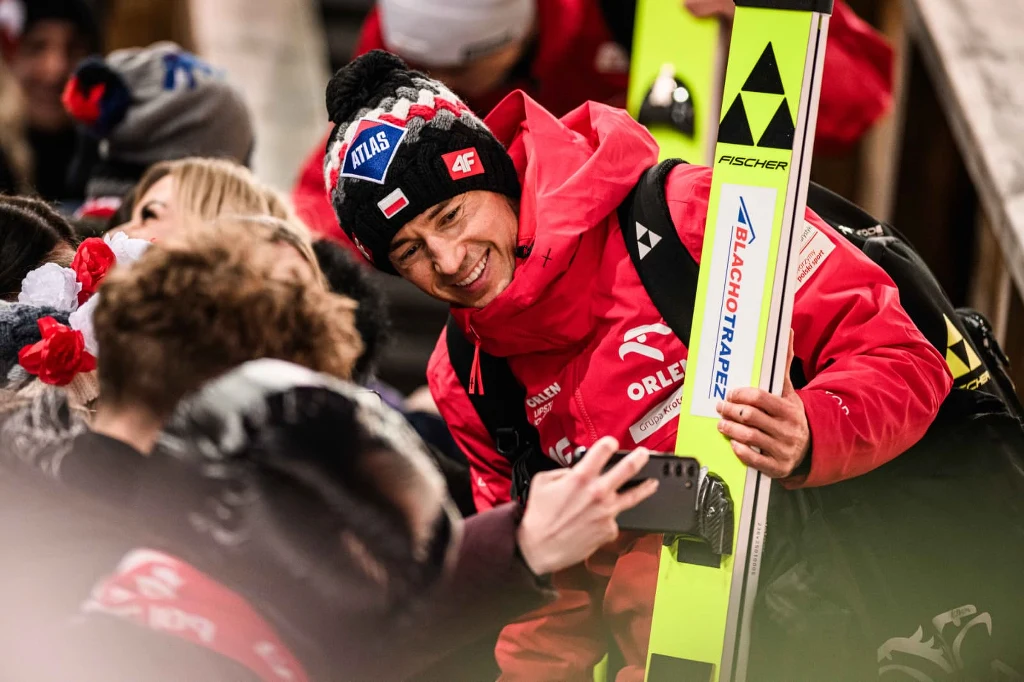 Kamil Stoch Uśmiechnięty skoczek narciarski w czerwonym kombinezonie trzyma narty i pozuje do selfie z kibicami. W tle znajdują się inne osoby ubrane w zimowe ubrania, a całość utrzymana jest w sportowej atmosferze.