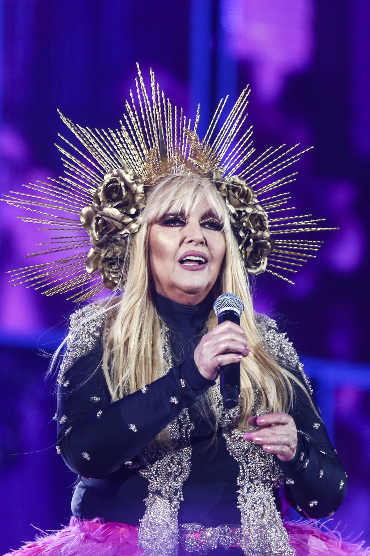 Maryla Rodowicz. Zakopane Sylwester Marzeń z Dwojką TVP Kobieta z długimi blond włosami w efektownym, ozdobnym kostiumie, trzymająca mikrofon, na głowie bogata korona z elementami przypominającymi promienie i złote kwiaty, tło utrzymane w fioletowych tonach oświetlenia scenicznego.