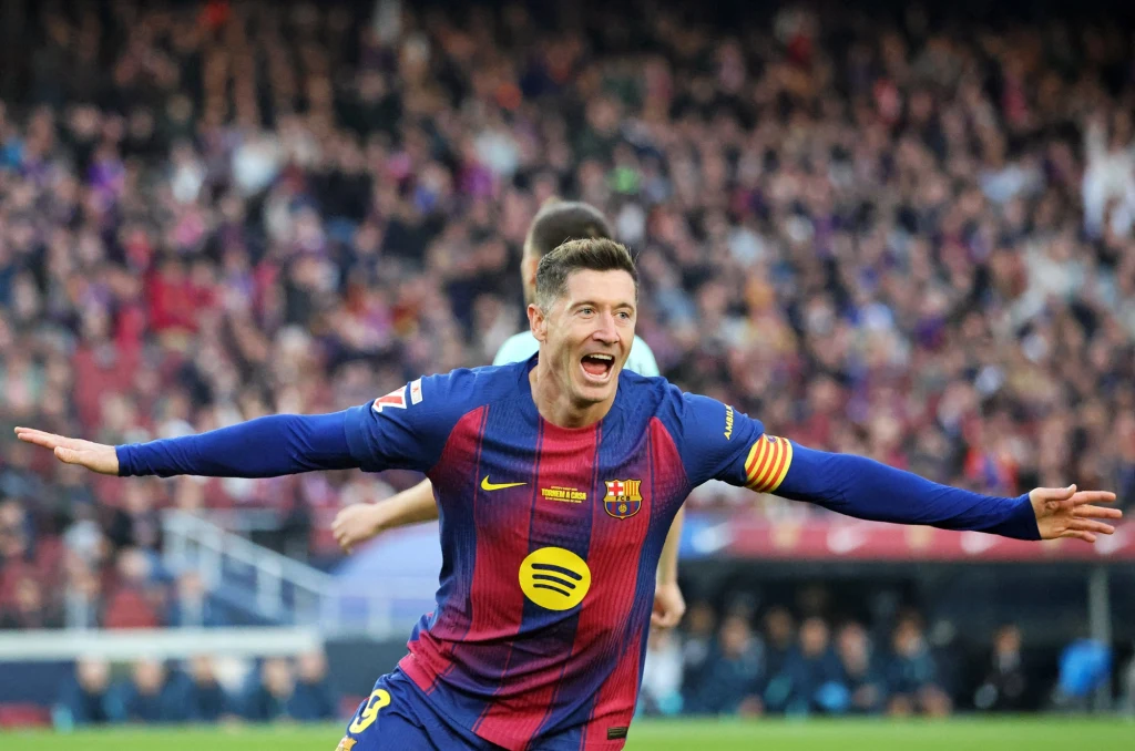 Robert Lewandowski w koszulce FC Barcelona podczas sezonu 2025/2026 Piłkarz Barcelony w pełnym stroju klubowym świętuje zdobycie bramki, rozkładając szeroko ręce. W tle rozmyta publiczność stadionu podczas meczu piłkarskiego.