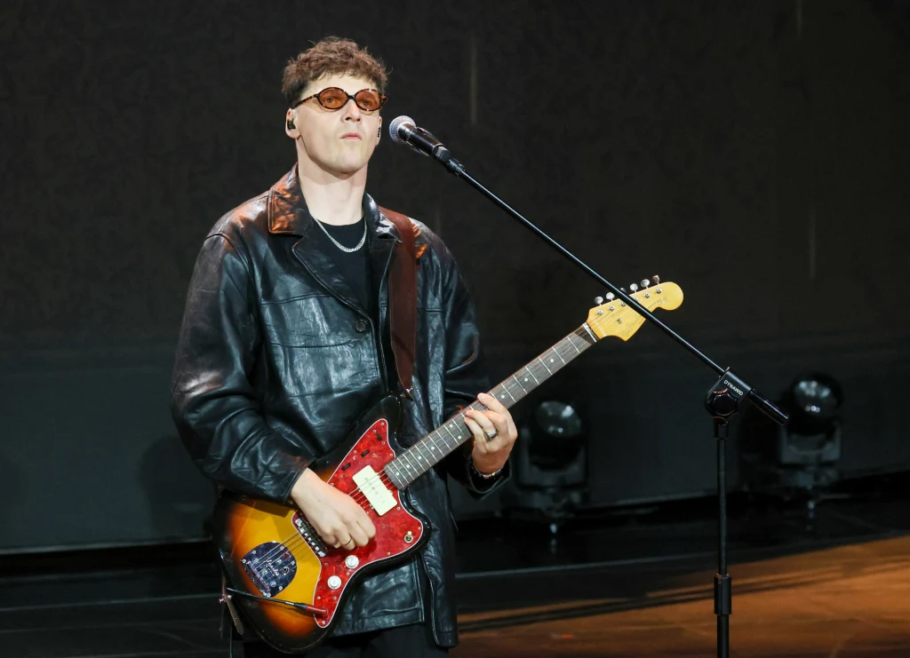 Muzyk w ciemnych okularach i czarnej skórzanej kurtce gra na elektrycznej gitarze na scenie, przed mikrofonem, na tle ciemnego oświetlenia.