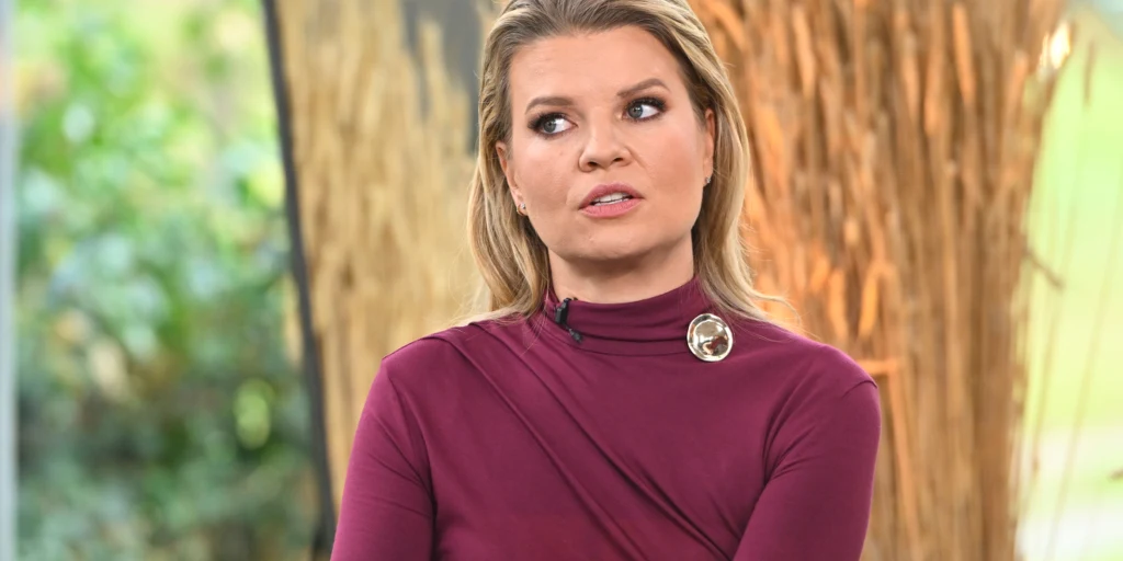 Kobieta o blond włosach w bordowej bluzce z ozdobną broszką, siedząca w jasnym wnętrzu z widocznymi elementami dekoracyjnymi w tle.