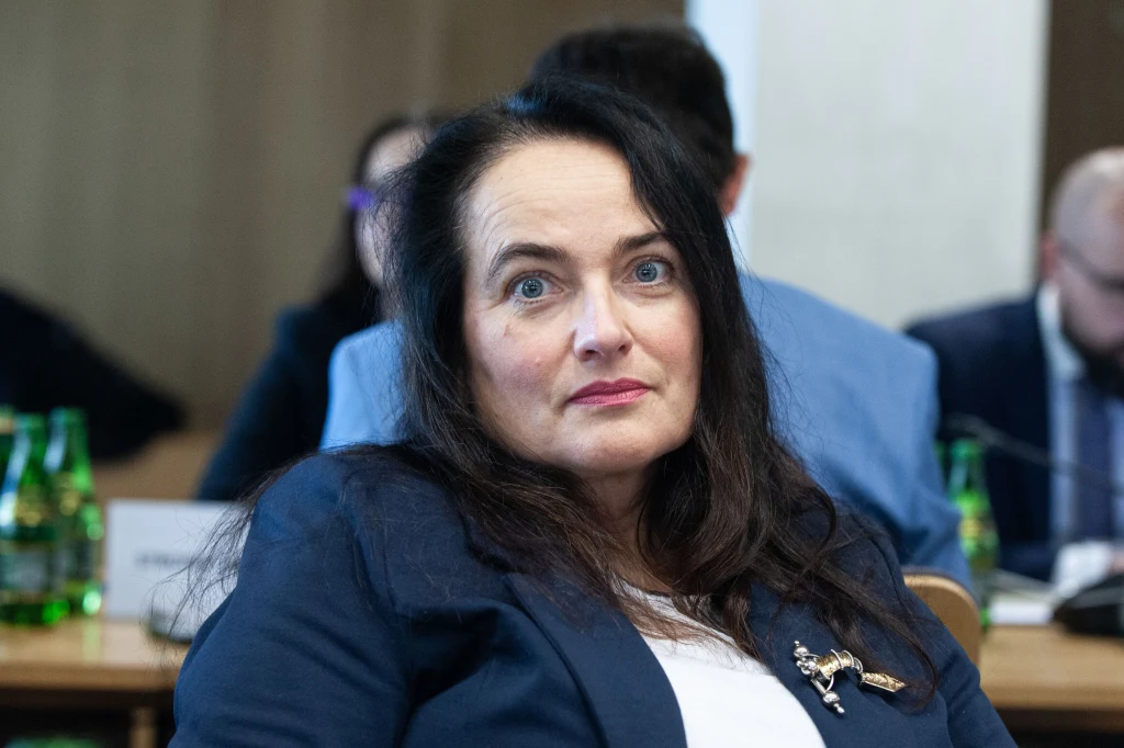 Kobieta o ciemnych włosach w eleganckiej marynarce siedzi przy stole w sali konferencyjnej, w tle widoczne osoby oraz butelki zielonej wody mineralnej.