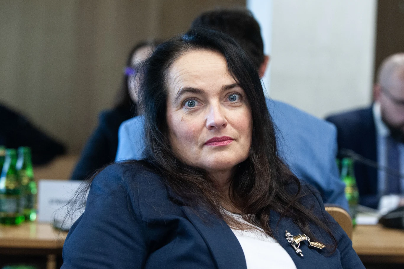 Anita Kucharska-Dziedzic Kobieta o ciemnych włosach w eleganckiej marynarce siedzi przy stole w sali konferencyjnej, w tle widoczne osoby oraz butelki zielonej wody mineralnej.