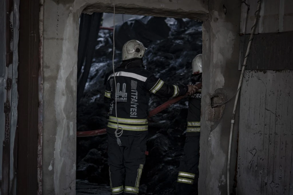 Dwóch strażaków ubranych w ciemne mundury i hełmy stoi na tle zniszczonego pomieszczenia, w którym widoczne są ślady po pożarze i pogorzelisko. Jeden z nich trzyma wąż pożarniczy, a pomiędzy nimi widać wejście do ciemnego wnętrza.