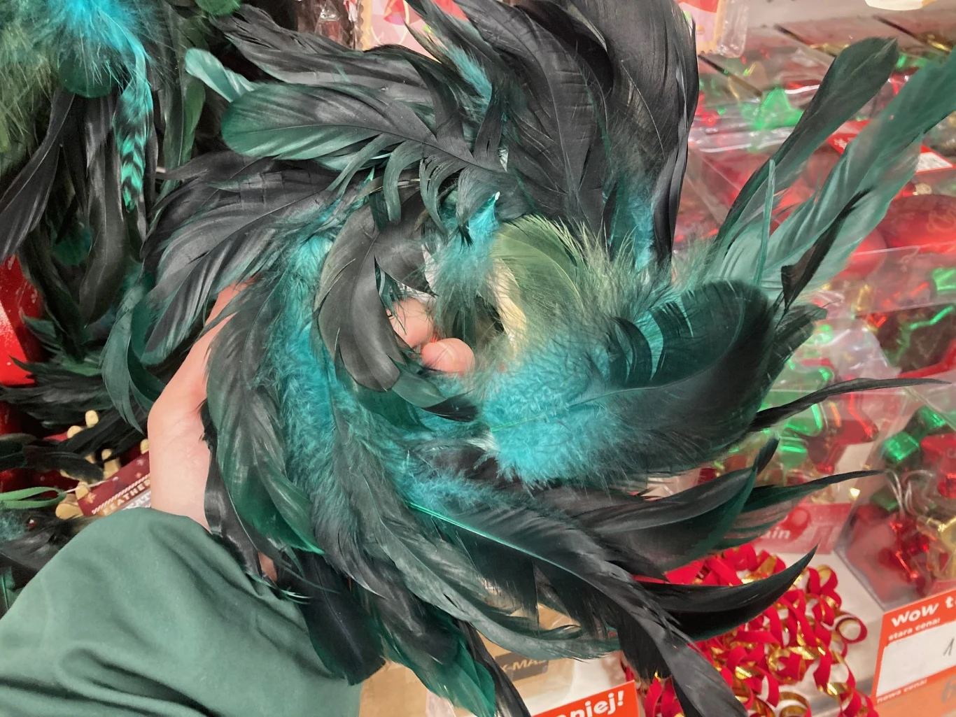 Ciemnozielone oraz turkusowe pióra trzymane w dłoni, w tle widoczne pudełka z ozdobami oraz czerwone wstążki.