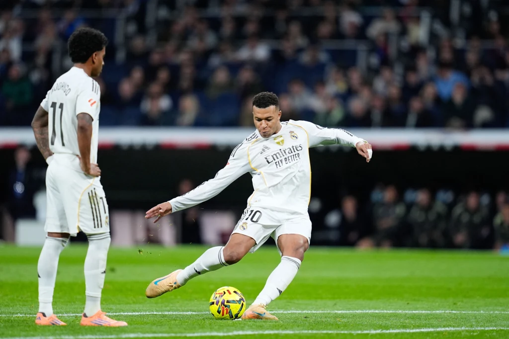 Dwóch piłkarzy w białych strojach Realu Madryt podczas meczu, jeden z nich wykonuje strzał w kierunku piłki, drugi obserwuje akcję stojąc obok. W tle widoczna publiczność na stadionie.