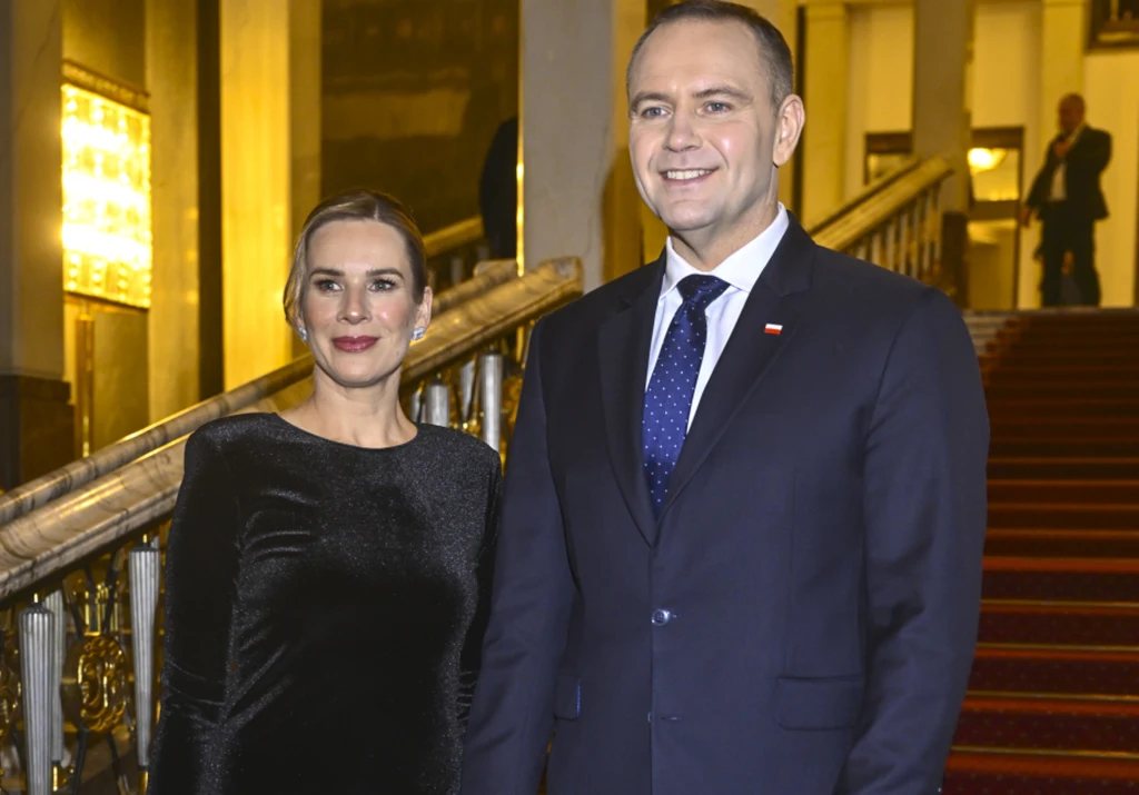Elegancko ubrana kobieta w czarnej sukni i mężczyzna w garniturze z niebieskim krawatem stoją razem w jasnym, reprezentacyjnym wnętrzu z czerwonym dywanem na schodach. Oboje uśmiechnięci, w tle widać elementy wystroju klasycznego budynku.