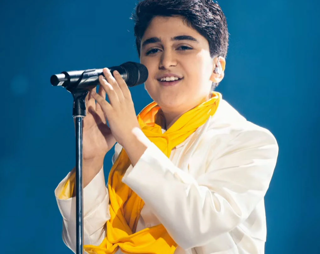Młody chłopak ubrany w biały garnitur z żółtym szalikiem śpiewa do mikrofonu na scenie, w tle jednolity niebieski kolor.