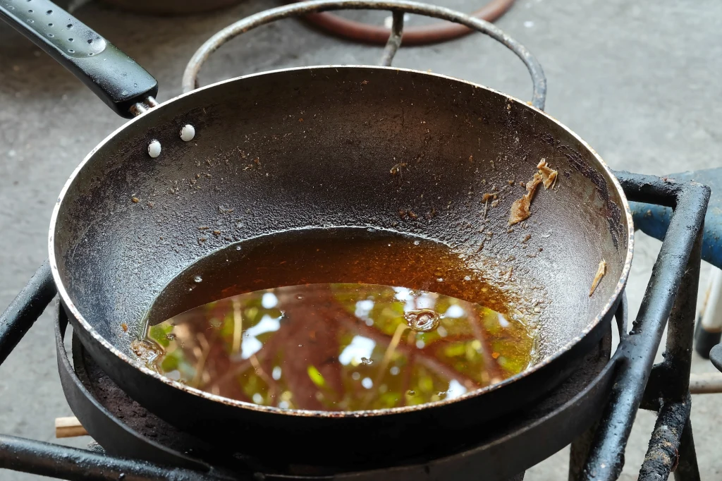 Nadmiar tłuszczu na patelni nie może być wylany do zlewu Stara, żelazna patelnia z resztkami używanego oleju i przypalonym tłuszczem, ustawiona na kuchence gazowej na zewnątrz, widoczne ślady użytkowania oraz odbicia otoczenia w tłuszczu.