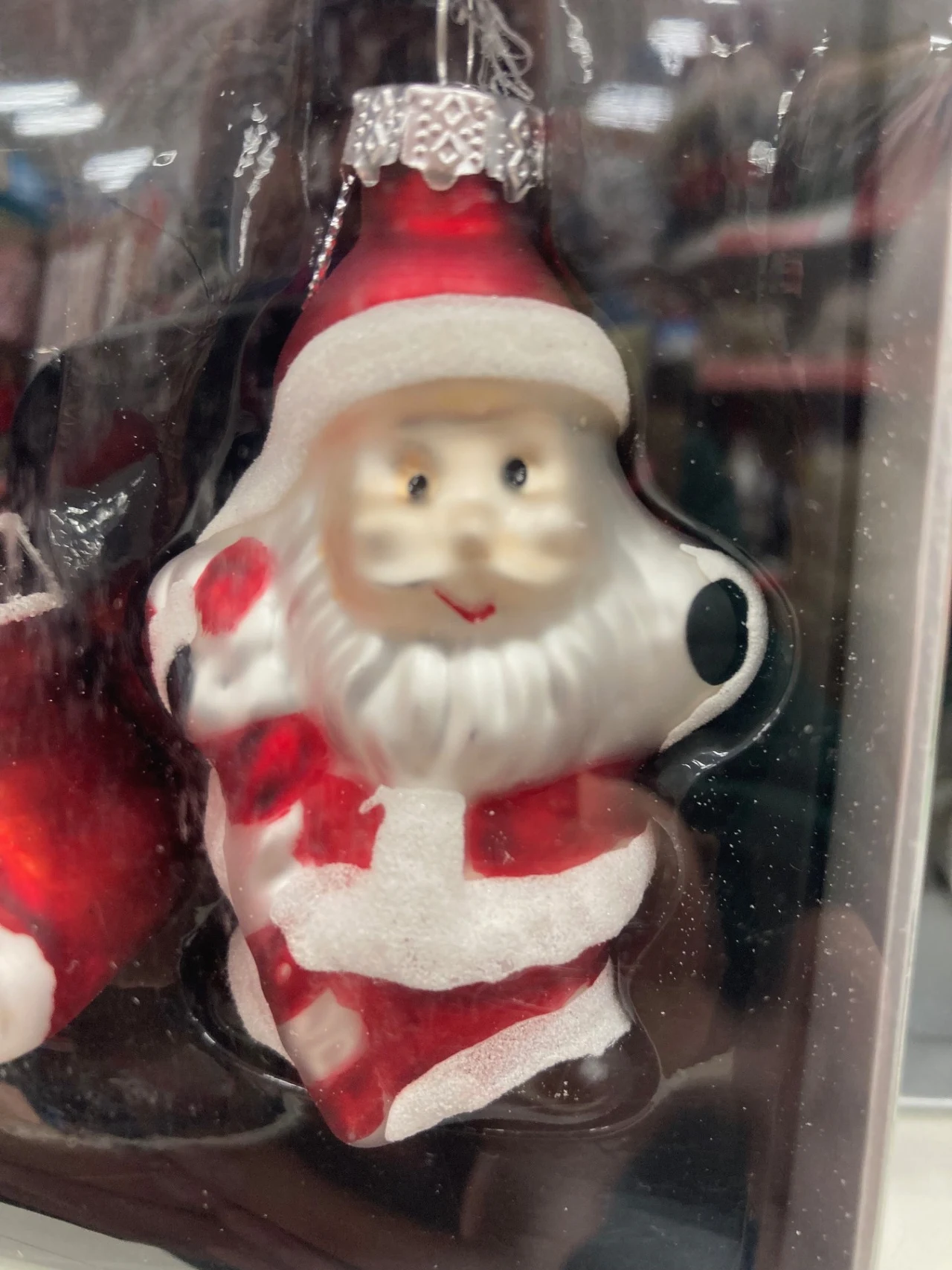 Szklana bombka choinkowa w kształcie Mikołaja z białą brodą i czerwonym strojem, trzymającego worek prezentów, ozdobiona srebrnym oczkiem do zawieszenia.