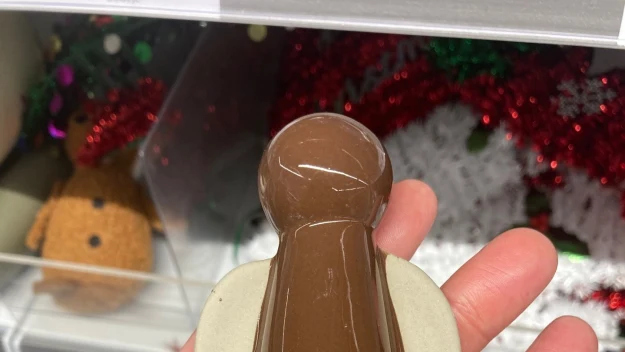 Figurka anioła z brązową szatą i jasnymi skrzydłami trzymana na dłoni, w tle półki sklepowe z ozdobami świątecznymi.