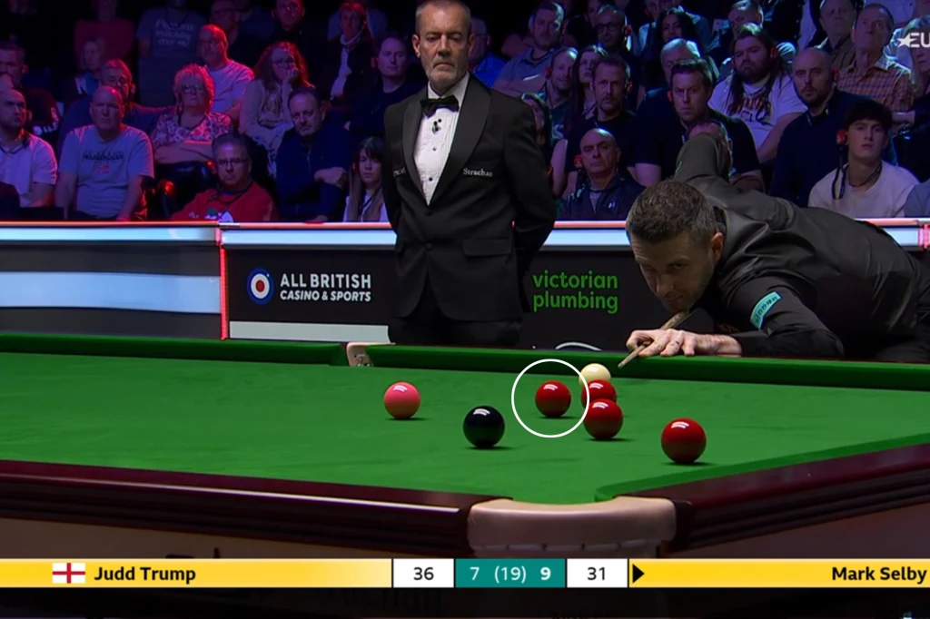 Potężny błąd Marka Selby'ego w 17. frejmie Zawodnik snookera wykonuje precyzyjne uderzenie nad zielonym stołem bilardowym, sędzia obserwuje przebieg rozgrywki, a w tle widoczna jest publiczność oraz podświetlane reklamy.