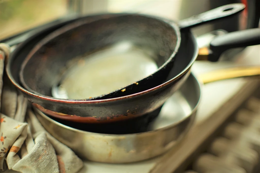 Ocet, sól, kwasek cytrynowy: niezawodne trio do czyszczenia garnków Stosy brudnych patelni ułożone na siebie na kuchennym blacie obok ściereczki, w tle rozmyte światło wpadające przez okno.