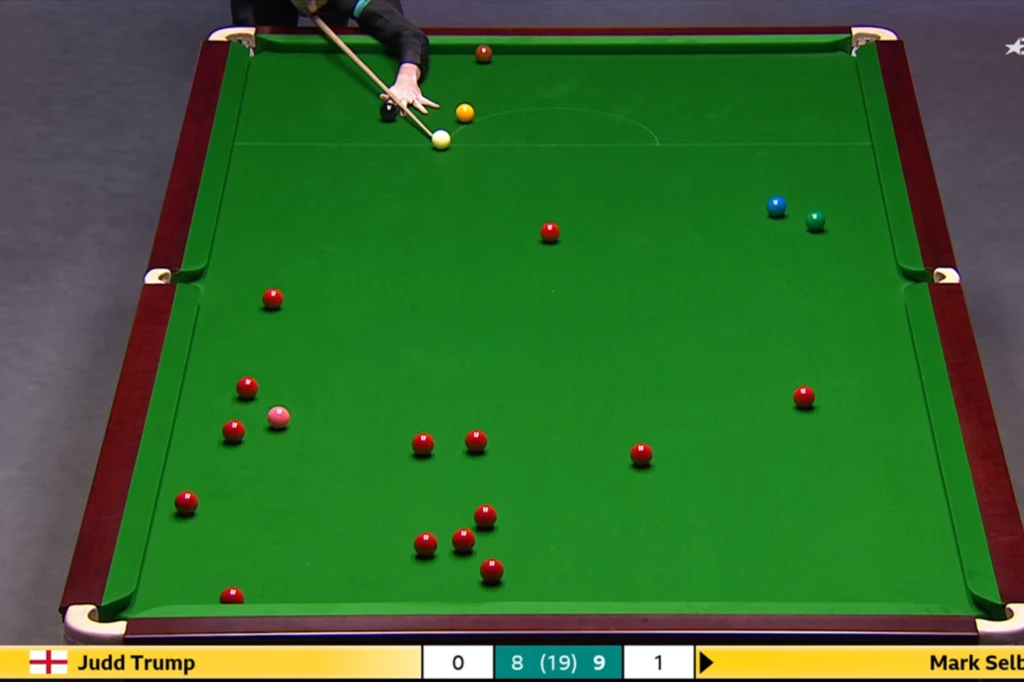 Ostatni frejm w wielkim finale UK Championship. Tu Mark Selby zaczął czyszczenie stołu Stół do snookera z rozstawionymi bilami, zawodnik przygotowujący się do uderzenia, widoczne dłonie oraz kij bilardowy, na dole plansza wyników z nazwiskami graczy i aktualnym wynikiem.