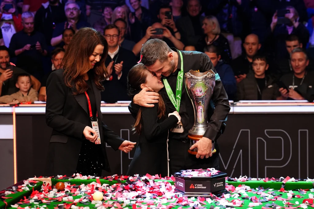 Mark Selby z żoną i córką - po triumfie w UK Championship Mężczyzna trzymający puchar i medale przytula młodą dziewczynę, obok kobieta uśmiecha się. Na stole widać konfetti i pudełko, w tle zgromadzeni kibice robią zdjęcia i biją brawo.