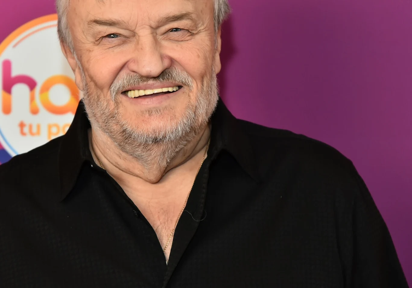 Krzysztof Cugowski jest jedną z najbardziej rozpoznawalnych postaci polskiej sceny Uśmiechnięty starszy mężczyzna o siwych włosach i zarostem ubrany w czarną koszulę pozuje na fioletowym tle z kolorem pomarańczowym widocznym w tle.