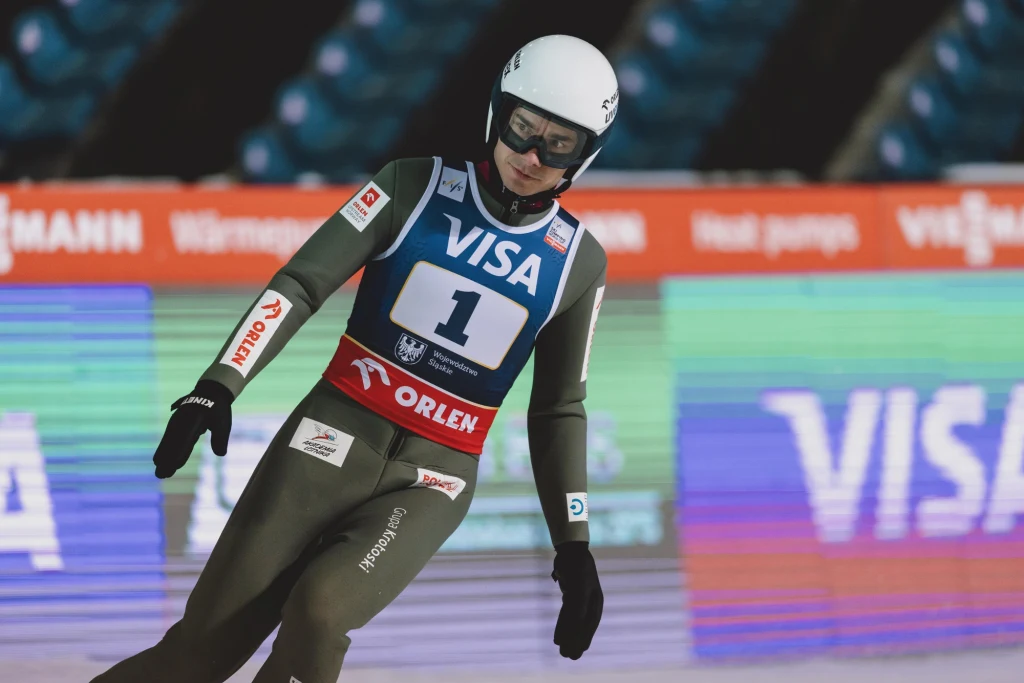 Jakub Wolny Zawodnik w kombinezonie narciarskim i kasku podczas lądowania po skoku narciarskim na tle kolorowych reklam i trybun stadionu.