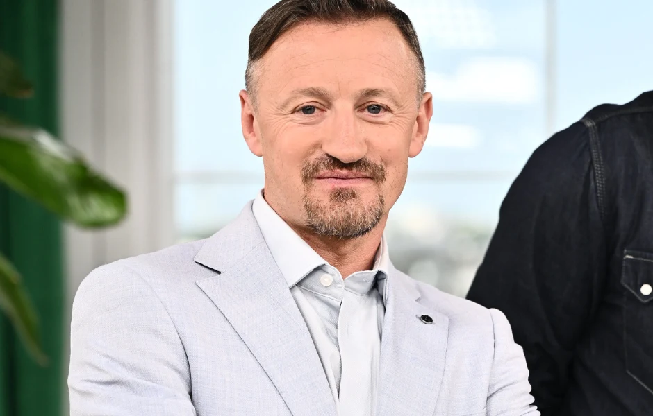 Mężczyzna w jasnym garniturze z białą koszulą i krawatem, z krótką brodą oraz włosami zaczesanymi na bok, uśmiecha się lekko w jasnym, nowoczesnym wnętrzu.