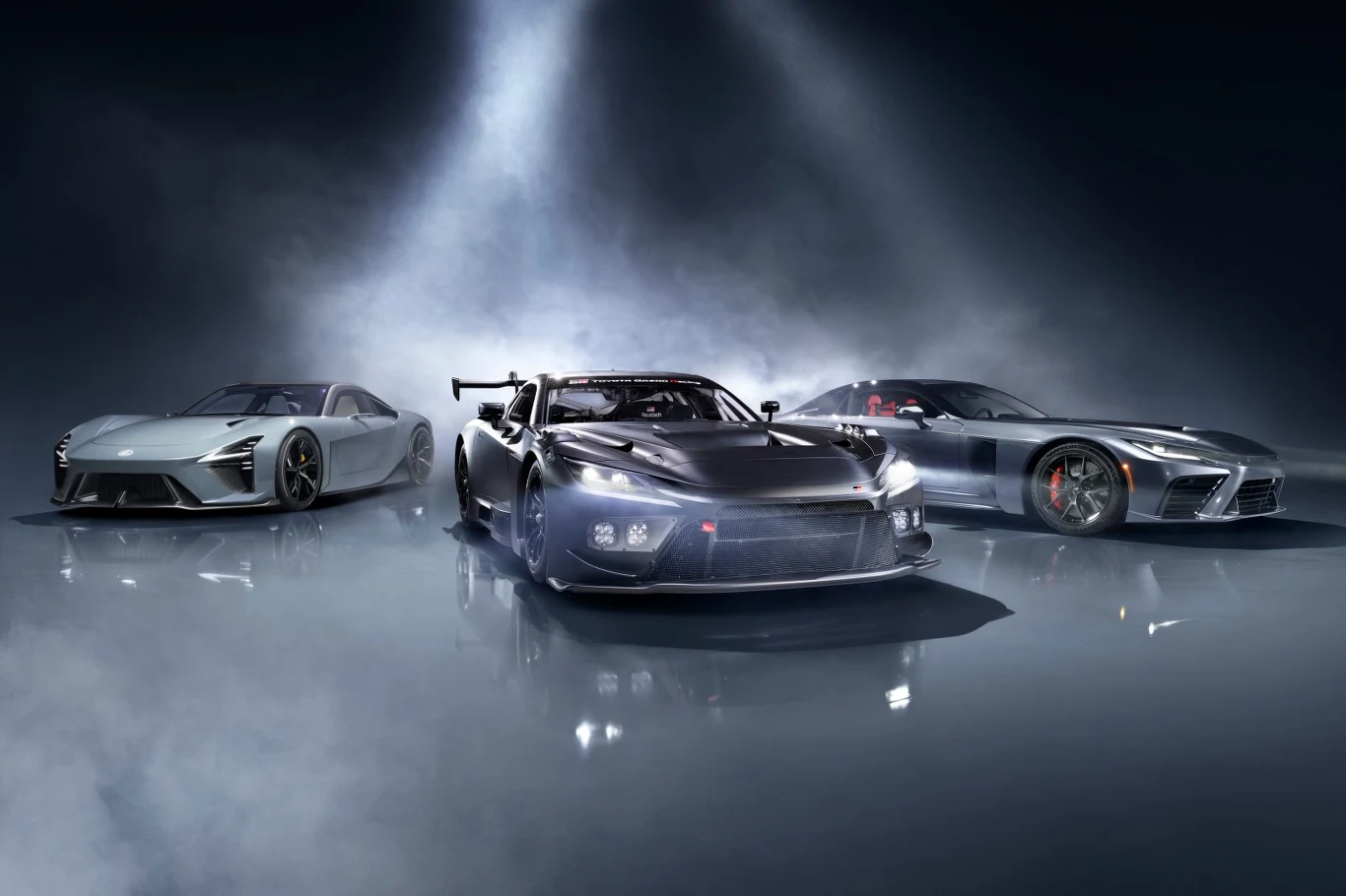 Trzy sportowe samochody ustawione obok siebie na połyskującej powierzchni, oświetlone reflektorem wydobywającym detale aerodynamicznych sylwetek i agresywnego designu każdego z pojazdów.