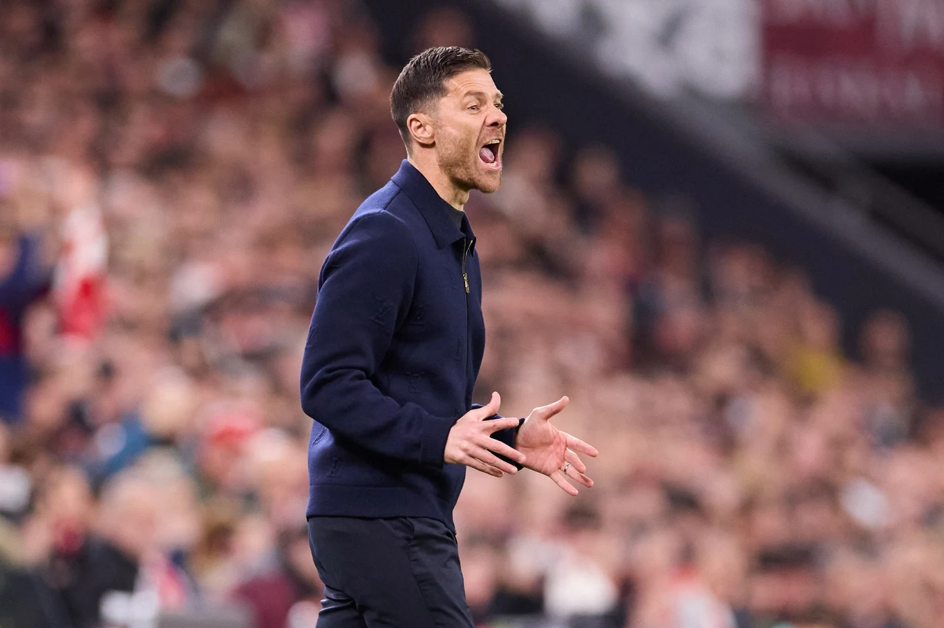 Xabi Alonso Mężczyzna ubrany w granatową marynarkę gestykuluje energicznie i krzyczy podczas wydarzenia sportowego na stadionie, w tle rozmyte sylwetki kibiców.