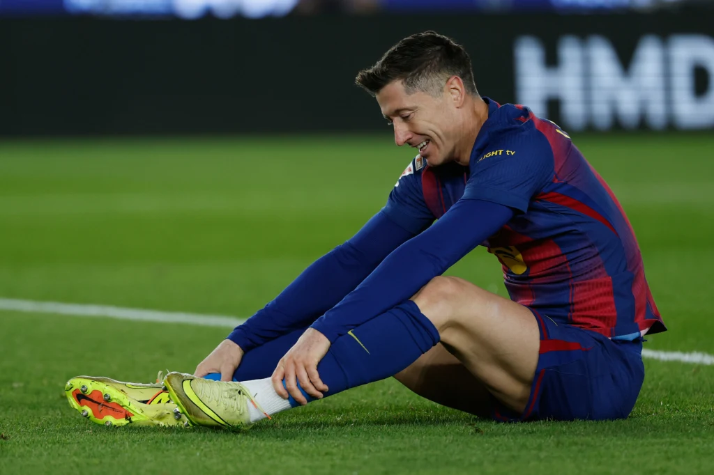 Robert Lewandowski Piłkarz w stroju meczowym siedzi na murawie boiska, trzymając się za lewe podudzie i sprawiając wrażenie lekko zmęczonego lub zaniepokojonego.