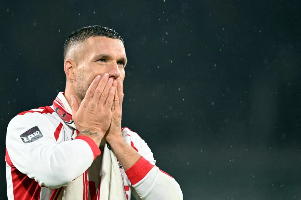 Piłkarz w biało-czerwonym stroju z dłońmi na twarzy, wyrażający silne emocje, na tle ciemnego, rozmytego tła.