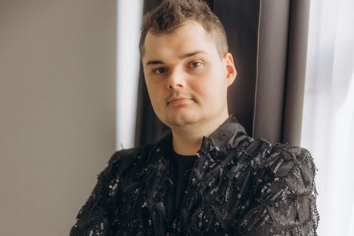 Michał Ławniczak w rozmowie z Interią wyjawia, jakie energie będą krążyć w 2026 roku Młody mężczyzna o jasnej cerze i ciemnych, krótkich włosach stoi z założonymi rękami przy oknie w jasnym pomieszczeniu, ubrany w czarną marynarkę z ozdobnymi wzorami.