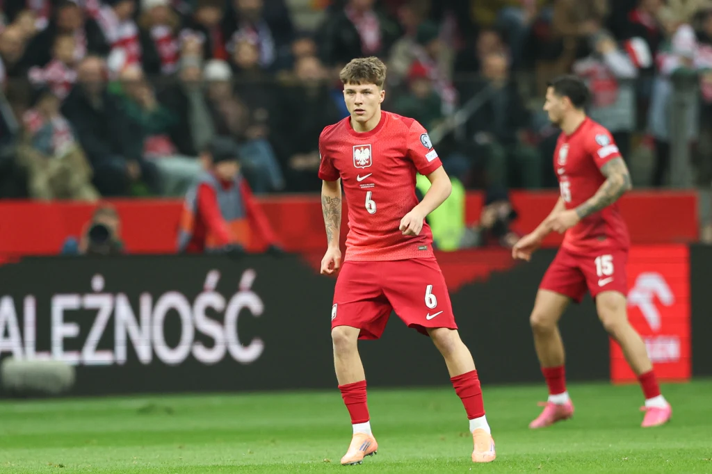 Polski piłkarz w czerwonym stroju narodowej reprezentacji na boisku podczas meczu, w tle rozmazani kibice i baner z napisem, drugi zawodnik w ruchu za główną postacią.