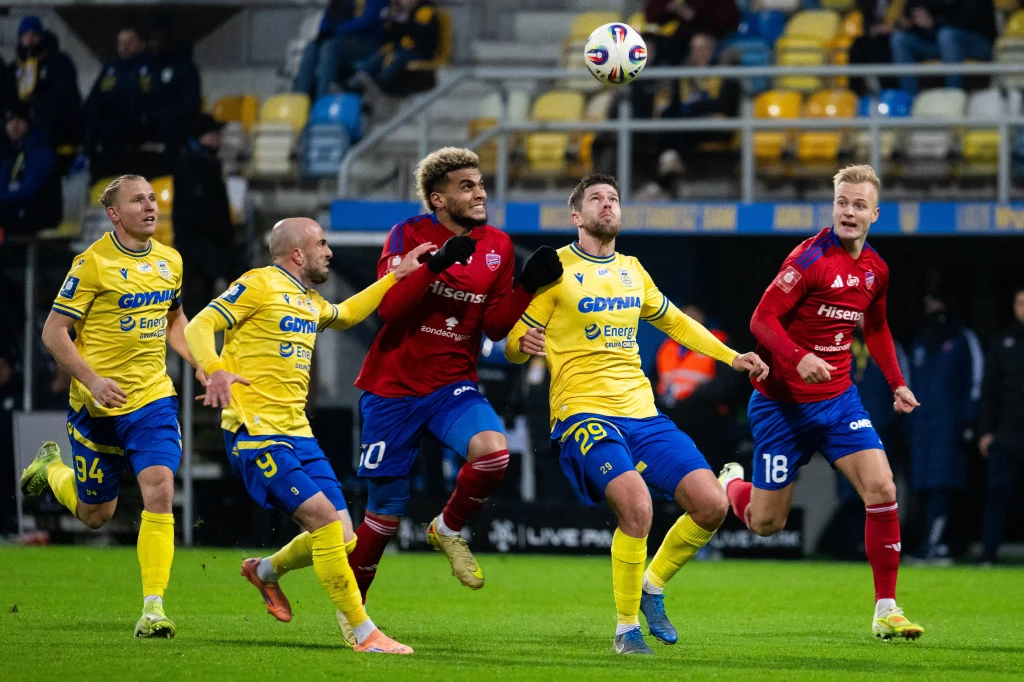 Pięciu piłkarzy w dynamicznej walce o piłkę podczas meczu piłkarskiego, trzech w żółto-niebieskich strojach i dwóch w czerwono-granatowych, w tle rozświetlone trybuny pełne pustych i częściowo zajętych krzeseł.