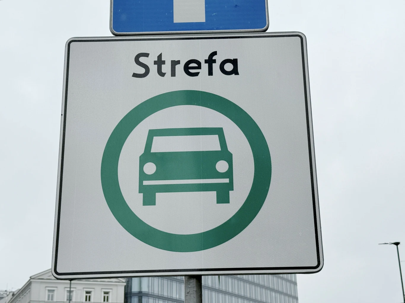 Ile należy zapłacić, żeby wjechać do SCT w Krakowie? Biały znak drogowy z napisem „Strefa” oraz zielonym symbolem samochodu w okręgu, umieszczony na tle miejskiej zabudowy i szarego nieba.
