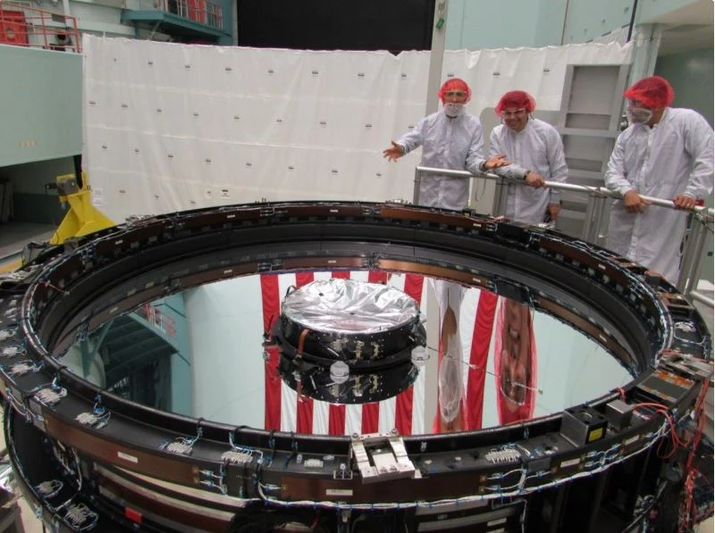 Inżynierowie w białych fartuchach i czerwonych czepkach ochronnych stoją przy dużym, okrągłym, metalicznym urządzeniu z odbijającą powierzchnią. Sprzęt wygląda na zaawansowany technologicznie, prawdopodobnie związany z badaniami naukowymi lub kosmicznymi.