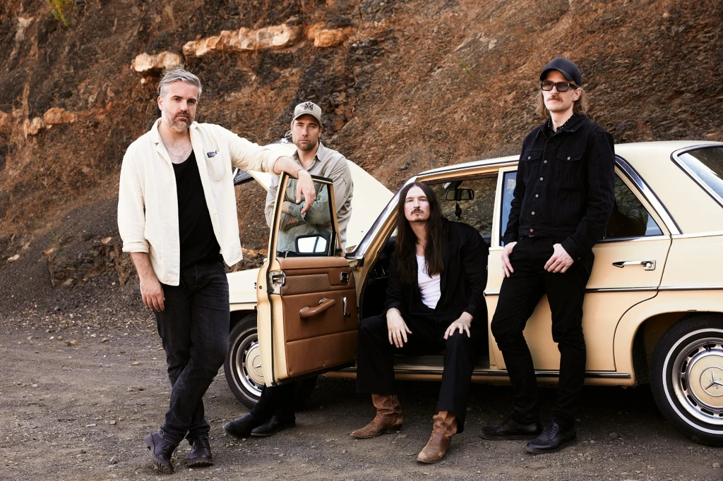 Kensington Czterech mężczyzn ubranych w swobodne ubrania stoi i siedzi przy jasnym klasycznym samochodzie na tle kamiennego zbocza, jeden z nich opiera się o otwarte drzwi auta, całość utrzymana w rockowym klimacie.