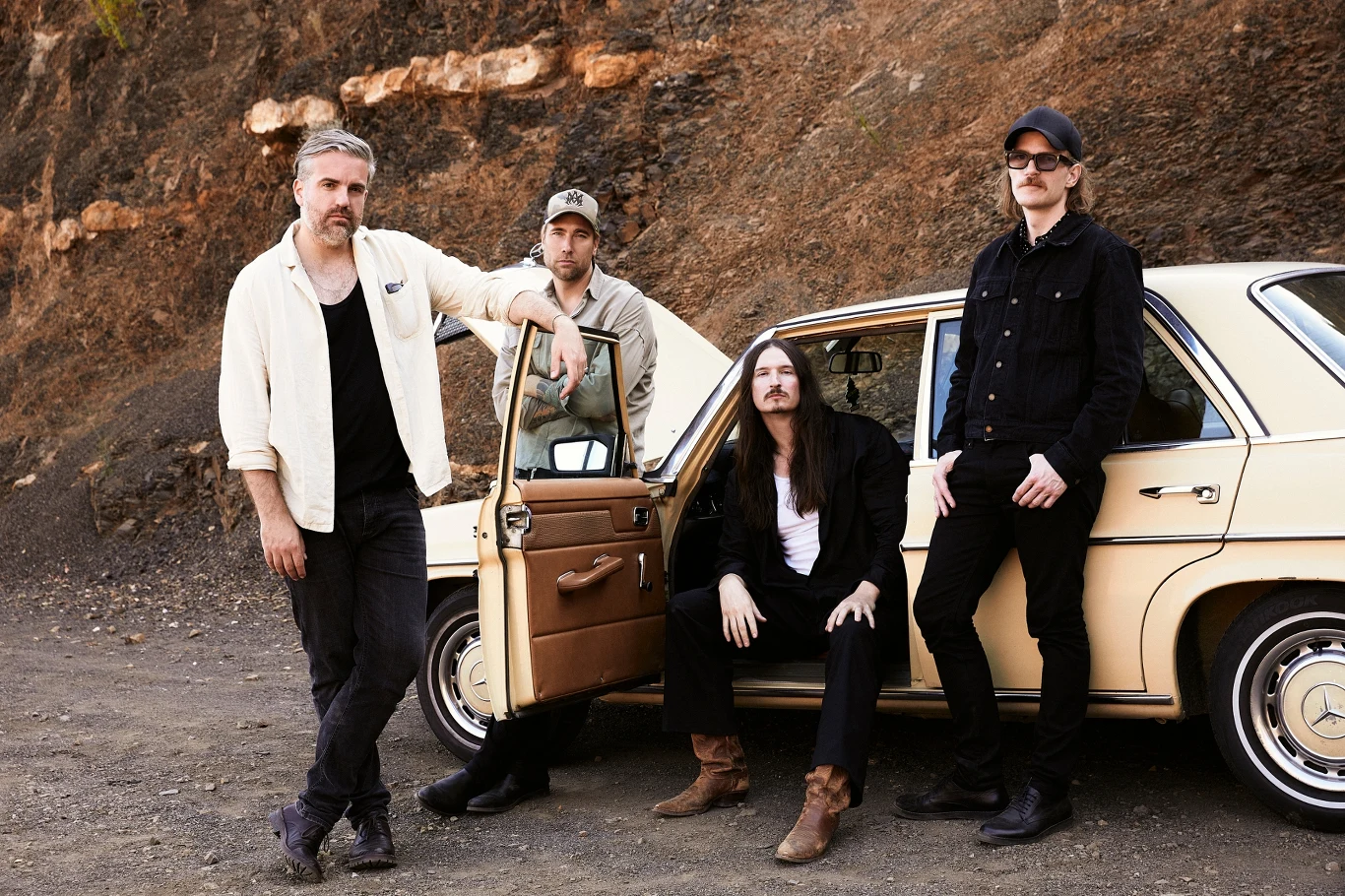 Czterech mężczyzn ubranych w swobodne ubrania stoi i siedzi przy jasnym klasycznym samochodzie na tle kamiennego zbocza, jeden z nich opiera się o otwarte drzwi auta, całość utrzymana w rockowym klimacie.
