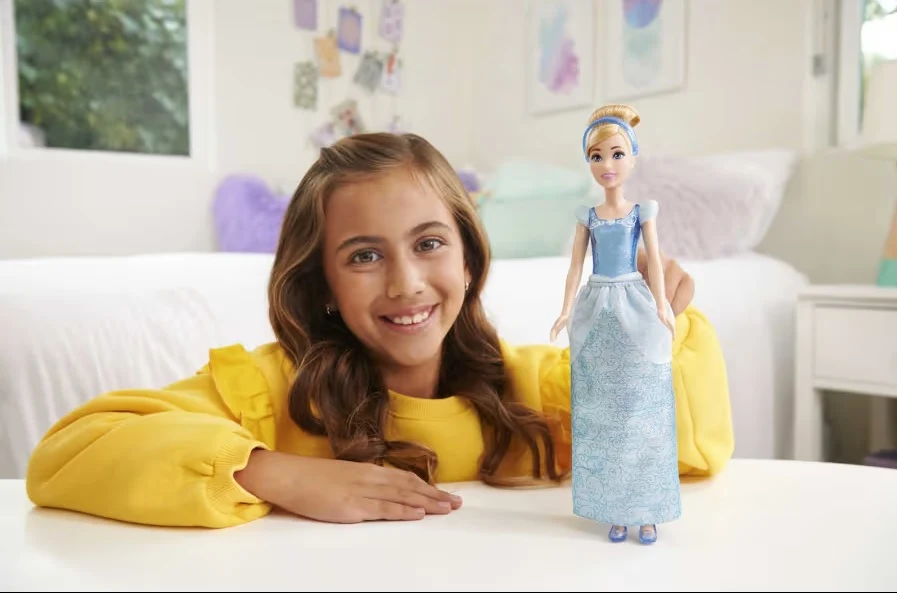 Współczesne zabawki sprawiają, że coraz częściej sami chcielibyśmy być znów dziećmi Dziewczynka z uśmiechem siedzi przy stole, trzymając w ręku lalkę ubraną w niebieską suknię. W tle jasny, przytulny pokój dziecięcy z łóżkiem, poduszkami oraz obrazkami na ścianie.