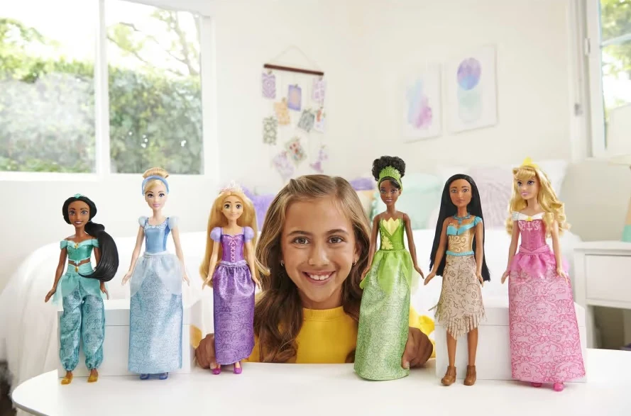 Bohaterki „Krainy lodu” to tylko jedna z propozycji disneyowskich lalek od Mattel Uśmiechnięta dziewczynka siedzi w jasnym pokoju przy stole, wokół niej ustawione są lalki przedstawiające księżniczki Disneya. Każda lalka ma kolorową suknię i długie włosy, a tło stanowi biały pokój z dekoracjami na ścianie.