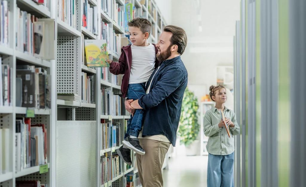 Ojciec trzymający syna na rękach wybiera dla niego książkę z półki w bibliotece, w tle kobieta trzymająca książkę i przyglądająca się z uśmiechem.
