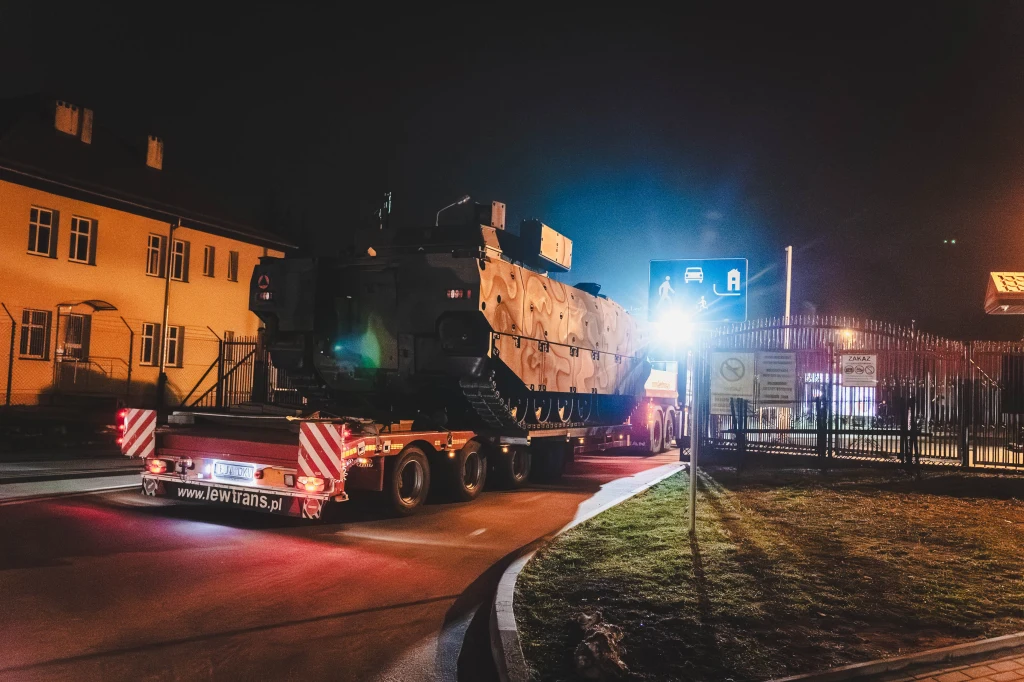 Ciężarówka niskopodwoziowa transportuje wojskowy pojazd opancerzony przez oświetloną ulicę w nocy. W tle widoczne budynki mieszkalne oraz ogrodzenie z bramą, całość otacza miejski krajobraz i sztuczne światła.