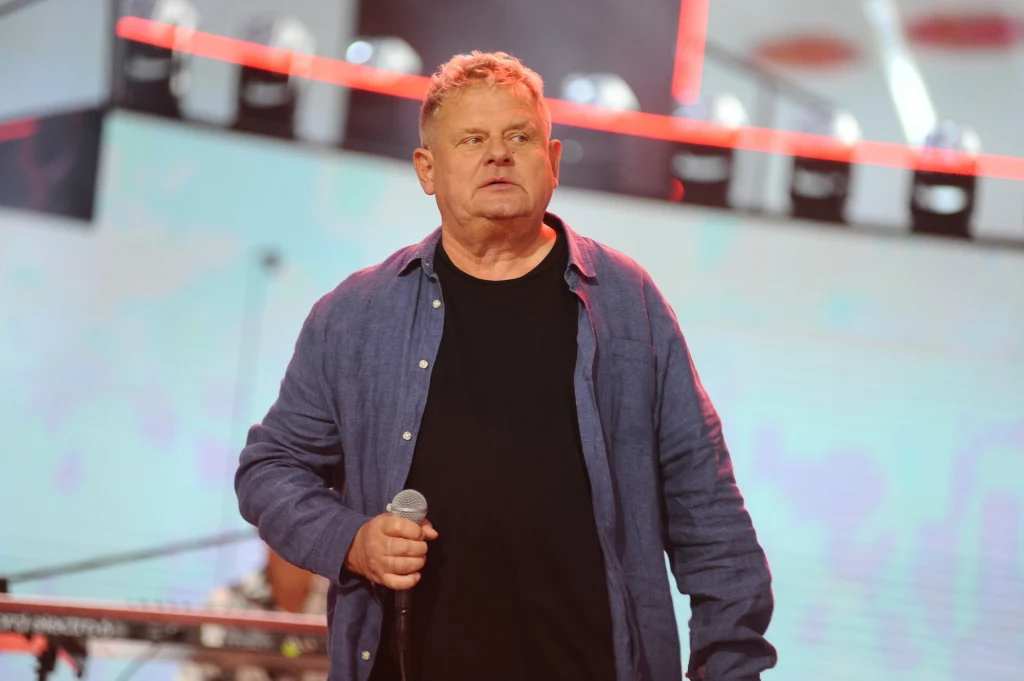 Mężczyzna w średnim wieku trzyma mikrofon, ubrany w niebieską koszulę i czarną koszulkę, stoi na scenie na tle rozświetlonego scenicznego tła, w pobliżu widoczne fragmenty klawiatury i sprzętu muzycznego.