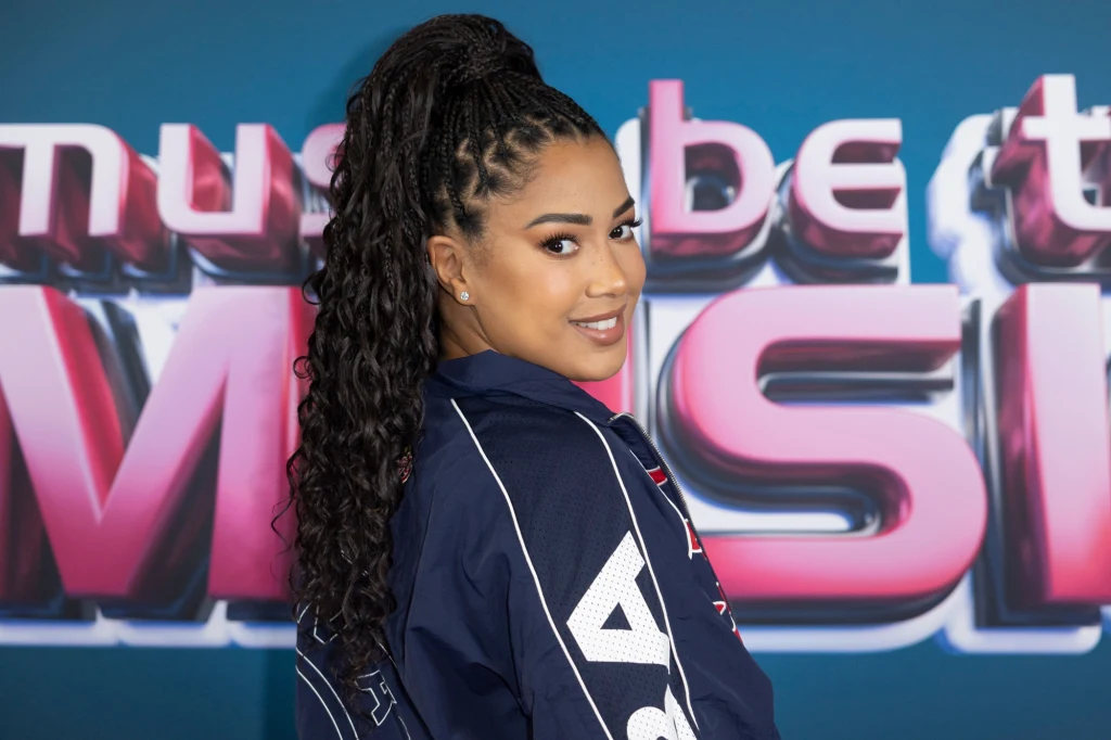 Patricia Kazadi Kobieta z upiętymi w loki włosami i uśmiechem na twarzy ubrana w granatową kurtkę pozuje na tle kolorowego napisu.