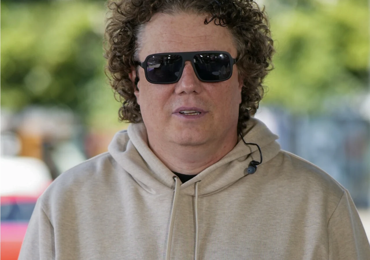 Sidney Polak Mężczyzna z kręconymi włosami, ubrany w jasną bluzę z kapturem i ciemne okulary przeciwsłoneczne, stoi na zewnątrz, na tle rozmytego zielonego otoczenia.