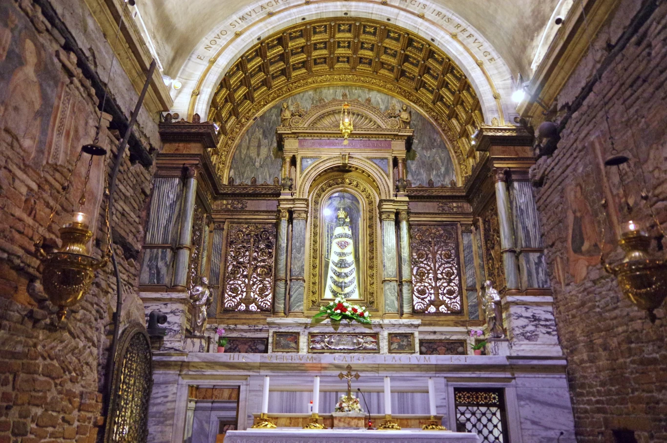 Wnętrze świętego domku we włoskim Lorento Bogato zdobiony ołtarz otoczony marmurowymi i złoconymi detalami, w centralnej części figura Madonny z Dzieciątkiem, całość pod arkadowym sklepieniem z freskami, ściany z cegły oraz liczne złocone elementy dekoracyjne.