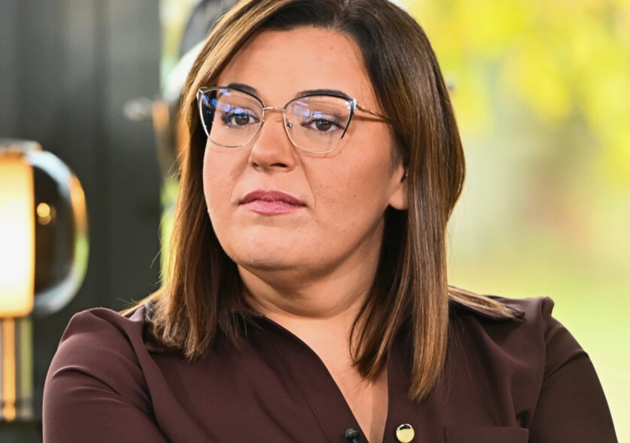 Basia z "Rolnik szuka żony" 12 Kobieta o ciemnych włosach w okularach, ubrana w ciemnobrązową koszulę, siedzi w jasnym wnętrzu z lampą w tle i zielonym krajobrazem za oknem.