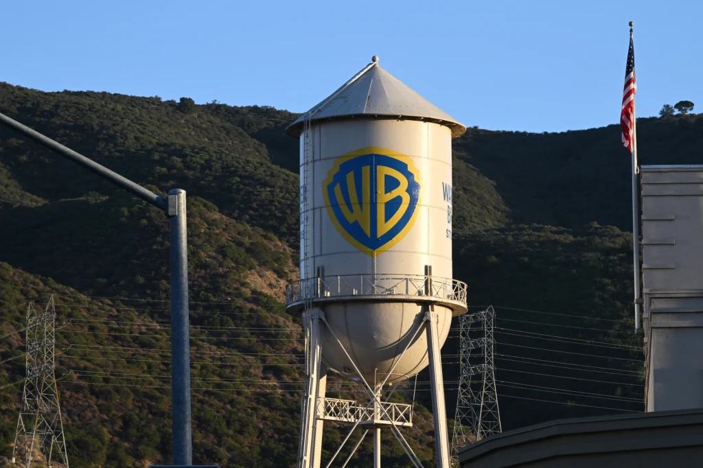 Duża wieża ciśnień z logotypem Warner Bros na tle gór i nieba, po lewej stronie widoczne słupy energetyczne, po prawej amerykańska flaga.