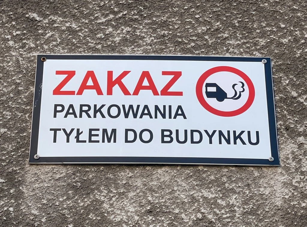 Takie tabliczki to w Polsce dość częsty widok. Tablica informacyjna z napisem zakazującym parkowania tyłem do budynku, czerwony znak przedstawiający samochód wydzielający spaliny, umieszczona na szarej ścianie.