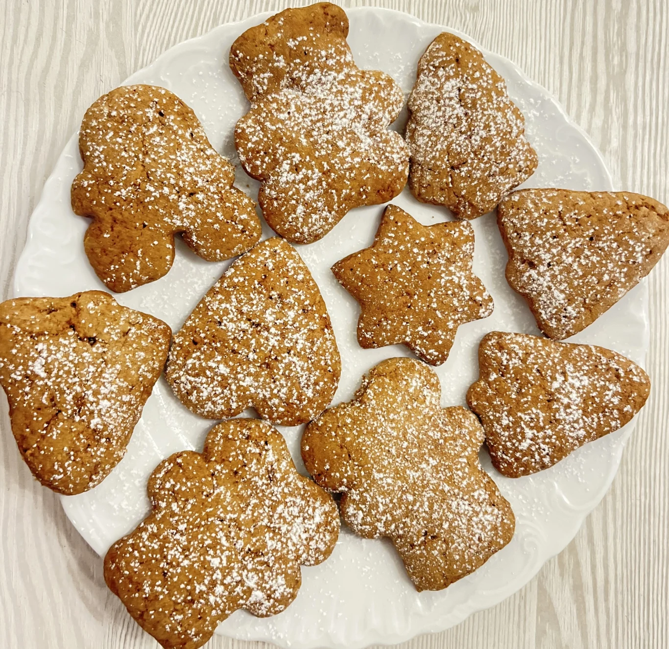 Pierniczki z przepisu mojej babci zawsze wychodzą pyszne - miękkie, puchate i aromatyczne Talerz z piernikami w kształcie choinek, gwiazdek i bałwanków, posypanymi delikatnie cukrem pudrem, ustawiony na jasnym, drewnianym tle.