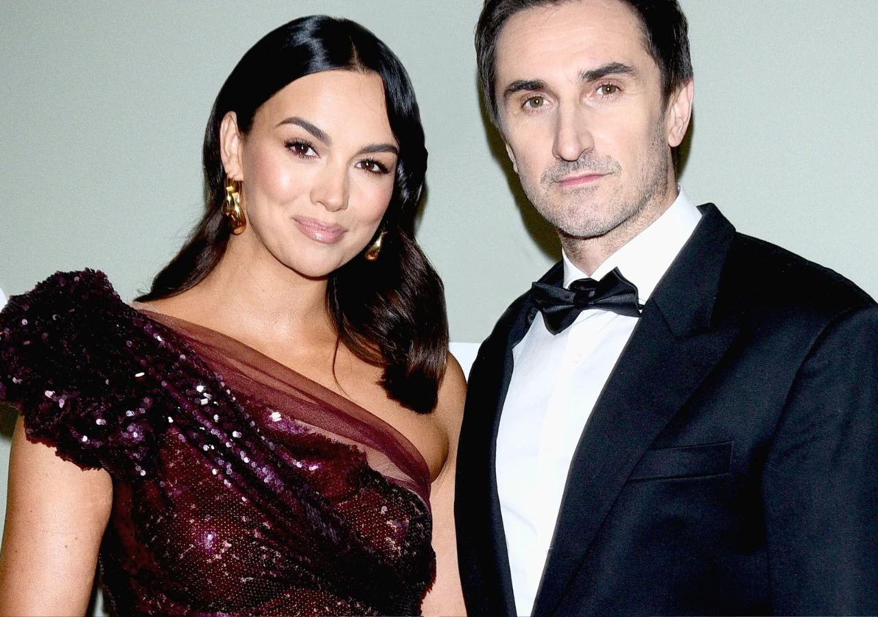 Paulina Krupińska, Sebastian Karpiel-Bułecka Elegancka kobieta w bordowej, połyskującej sukni i mężczyzna w czarnym smokingu z muszką, stoją razem pozując do zdjęcia, oboje uśmiechnięci, tło neutralne.