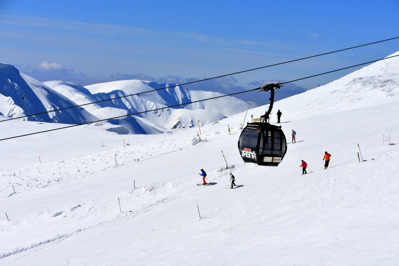 Ośnieżone stoki górskie z narciarzami korzystającymi z tras zjazdowych, nad którymi przejeżdża gondolowa kolejka linowa, w tle majestatyczne szczyty gór oraz błękitne niebo.