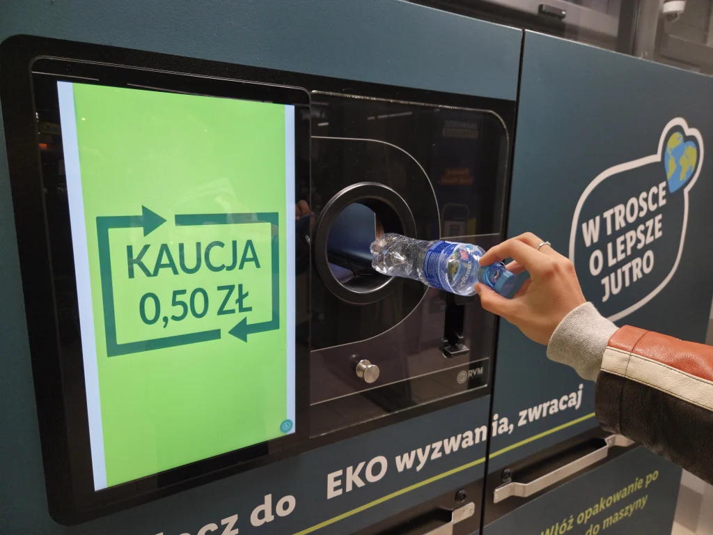 Czy za zwrot puszek i butelek z kaucją można zażądać gotówki? Ręka osoby wkłada plastikową butelkę do automatu recyklingowego, na ekranie wyświetla się informacja o zwrocie kaucji w wysokości 0,50 zł, po prawej stronie znajduje się napis W trosce o lepsze jutro.